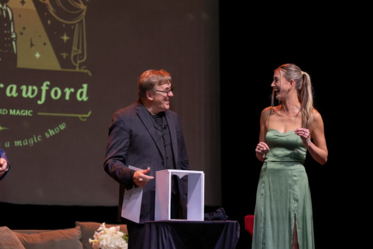 Rich Crawford laughing with a guest on stage during a corporate magic show, with the Crawford Magic logo projected behind them