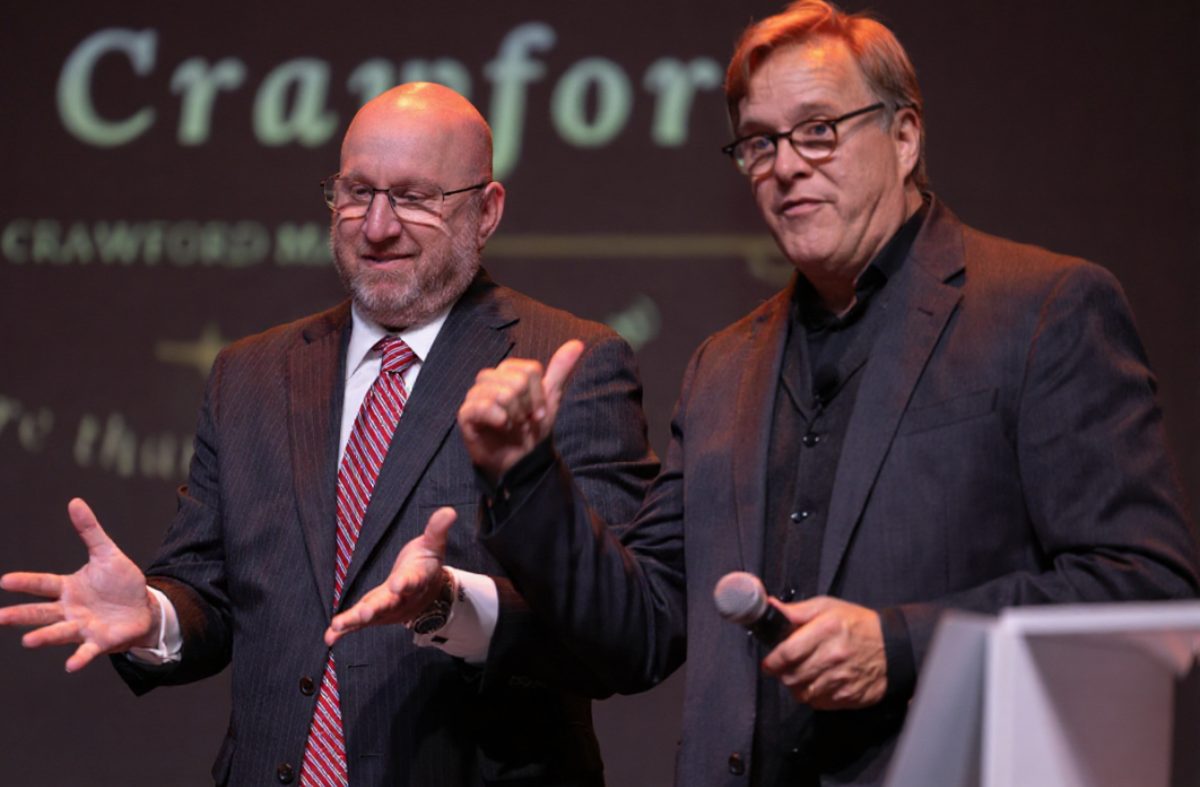 Rich Crawford on stage with a guest during a corporate magic show, with the Crawford Magic logo projected behind them